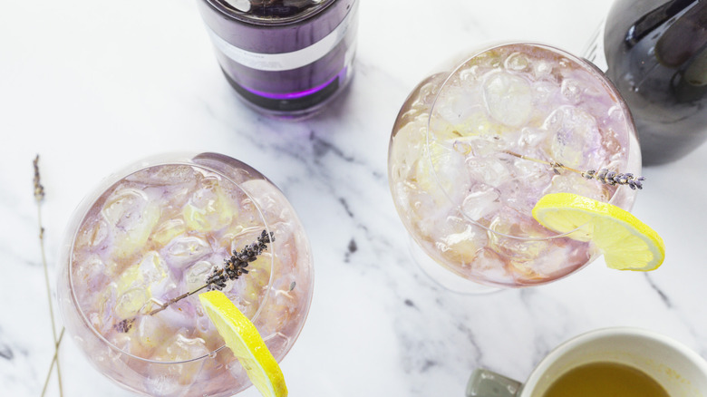 Overhead view of two glasses of layered lavender spritz cocktail, with gin and prosecco in background.