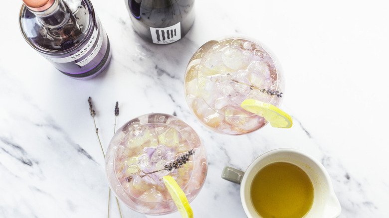 Overhead view of two glasses of layered lavender spritz cocktail, with gin and prosecco in background.