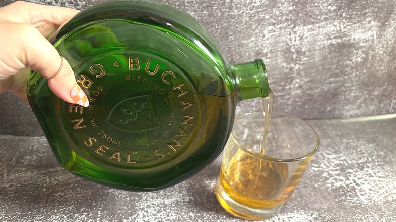 Hand pouring whisky from green bottle of Buchanan's Green Seal into whisky glass, all on black background