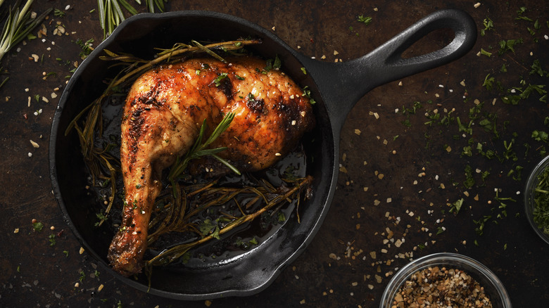 Roast chicken leg in a small cast iron skillet with herbs on a table scattered with various seasonings