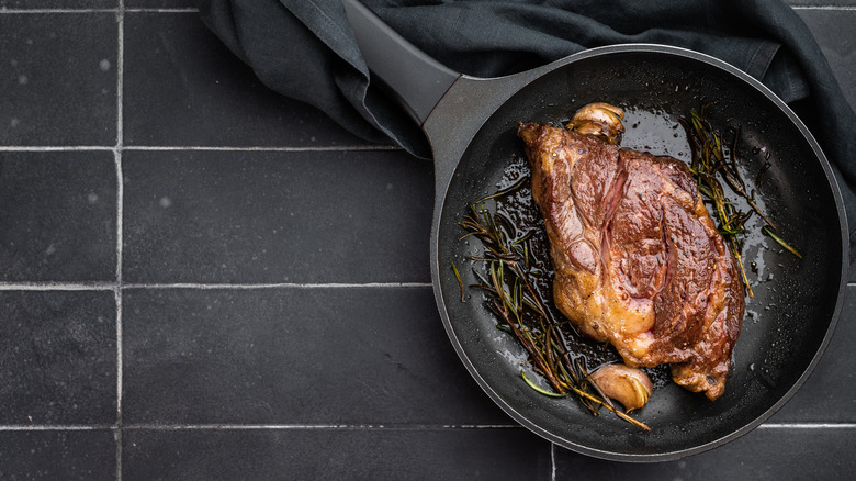 Pan seared chuck eye steak with a couple of cloves of garlic and herbs in a black pan on a black background.