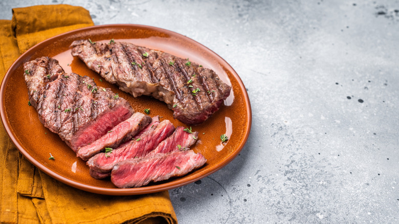 Two grilled flat iron steaks, one has been partially sliced, on a golden plate setting atop a golden linen napkin and gray background.