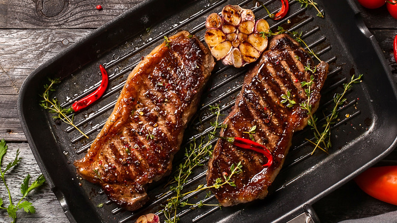 Two New York strip steaks on a griller pan with a head of garlic and rosemary, all sitting on a raw wooden table with tomatoes and peppers scattered around.