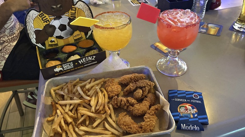 A tray of Buffalo Wild Wings chicken wings and fries in front of sauces and two cocktails