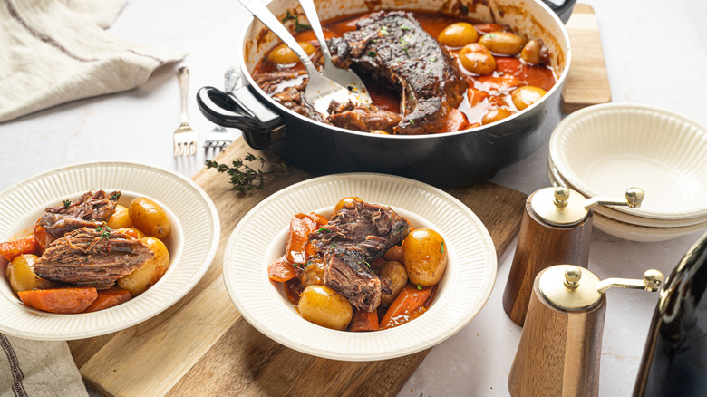 a pot of pot roast and plates on a wooden cutting board