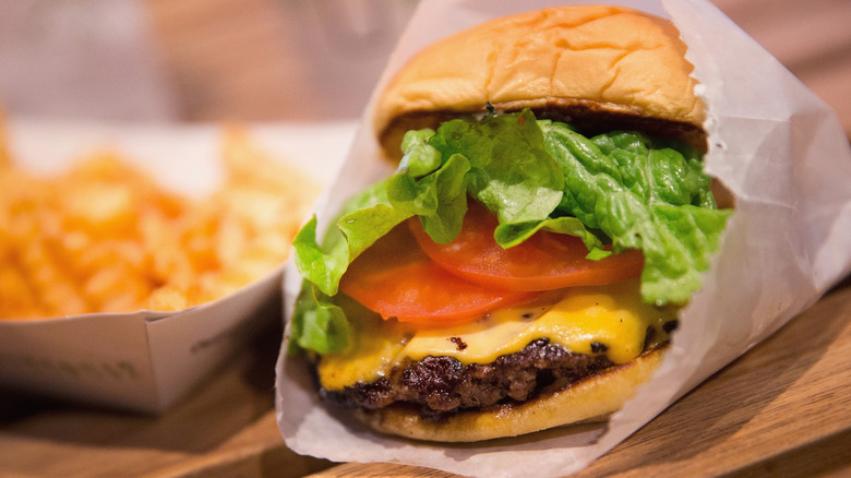 A cheeseburger with lettuce and tomato in a white paper bag next to a paper container of French fries
