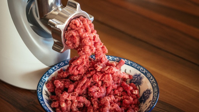 An electric meat grinder at home, with ground beef falling from it into a blue and white bowl