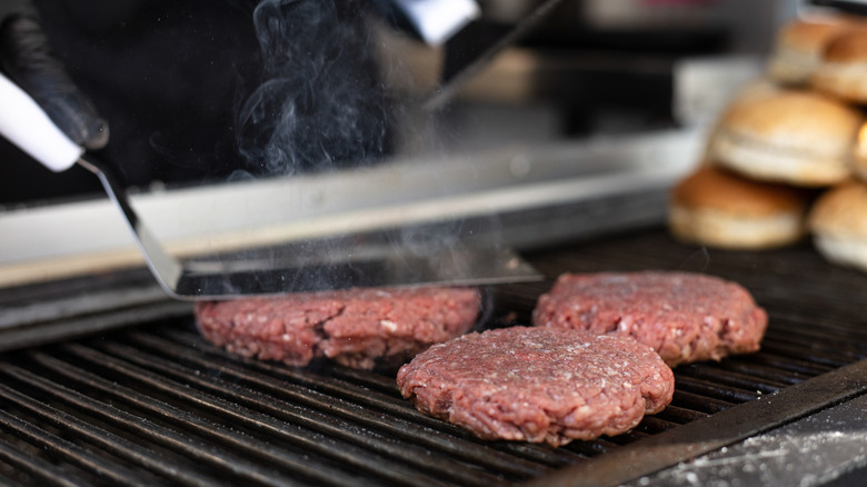 A cook presses a burger on the grill with a wide, stainless-steel spatula