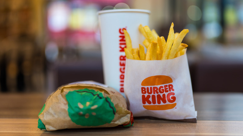 A combo meal from Burger King featuring a sandwich wrapped in paper, and envelope of french fries, and a drink sitting on a table