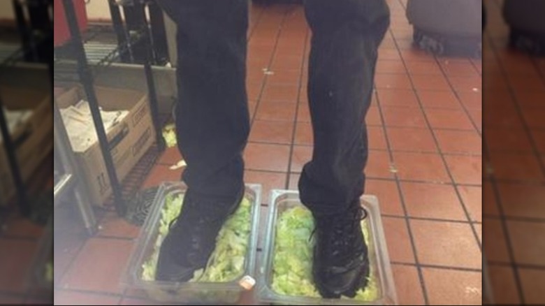 A pair of legs and black pants and black shoes standing in two plastic bins full of lettuce on a brown tile restaurant floor