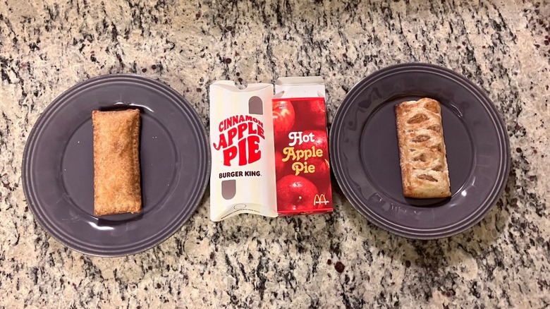 Burger King's cinnamon apple pie sits on a purple plate on the far left and McDonald's baked apple pie sits on a purple plate on the far right with each pie's sleeve between the plates