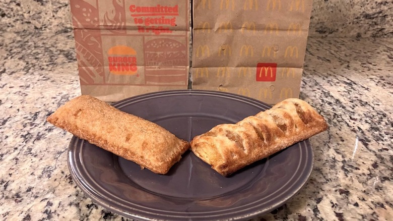 Burger King's cinnamon apple pie sits on the left side of a purple plate with the Burger King bag behind it and McDonald's baked apple pie sits on the right side of the purple plate with the McDonald's bag behind it