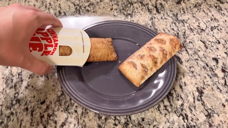 A hand shakes Burger King's cinnamon apple pie from its sleeve and onto a purple plate next to McDonald's baked apple pie