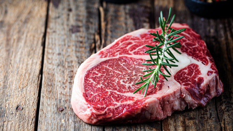 A ribeye steak sitting on a wooden table with a sprig of rosemary upon it