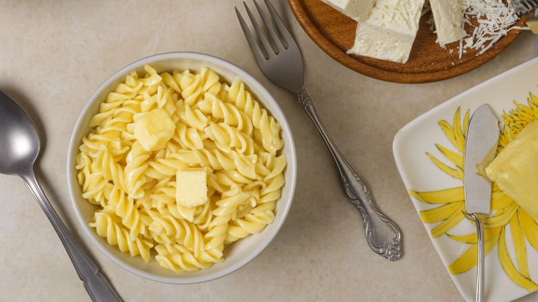 A white bowl of buttered noodles next to bowls of pasta, cheese, and butter