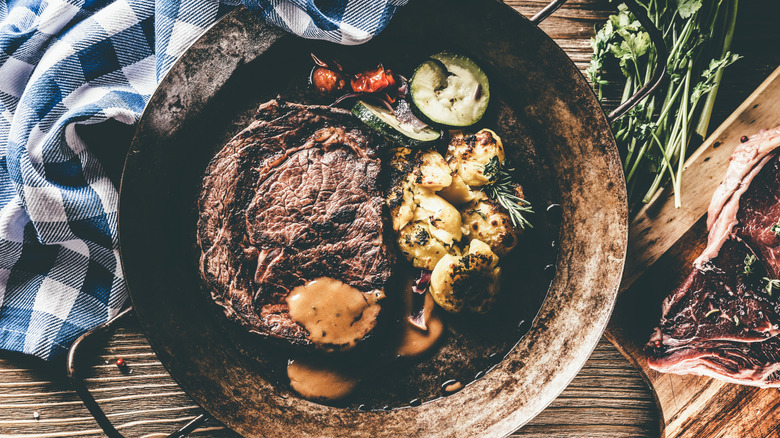 Steak rests in a rustic pan with sauce, garlic, herbs and vegetables