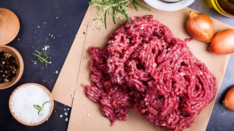 raw ground beef on butcher paper surrounded by salt, pepper, onions, and oil