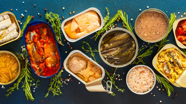 A variety of open tins of seafood surrounded by fresh herbs and peppercorns.