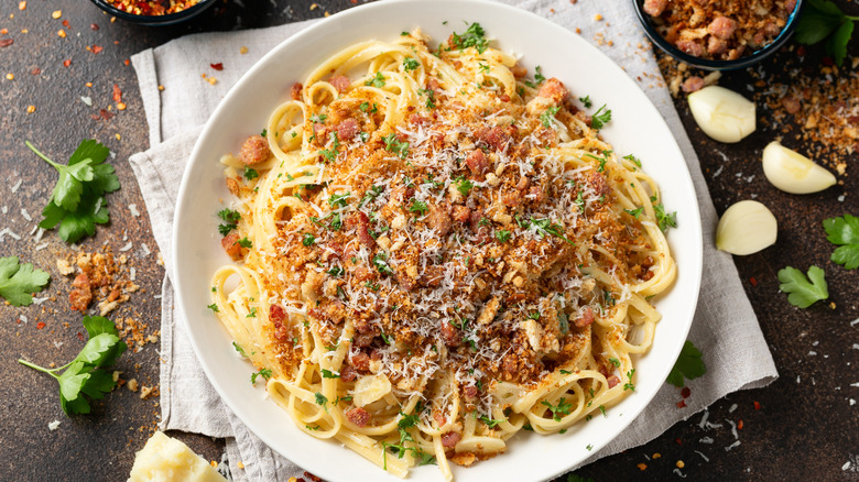 Plate of spaghetti topped with breadcrumbs and herbs next to bowl of breadcrumbs, spices, and herbs