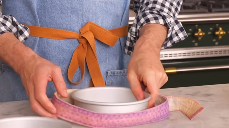 A person in a denim apron with a brown tie wraps a cake pan in a cake strip against a marble counter