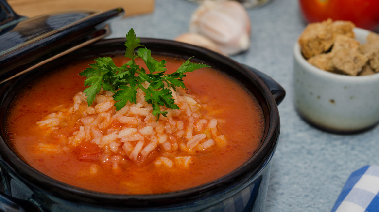 a small crock of tomato rice soup topped with a sprig of parsley with garlic and croutons beside it