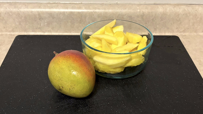 A fresh mango alongside a bowl of mango slices on a black cutting board.