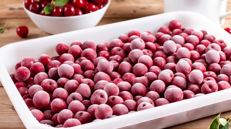 A plastic storage container full of frost covered Frozen cherries, with a bowl of fresh cherries on a table behind them