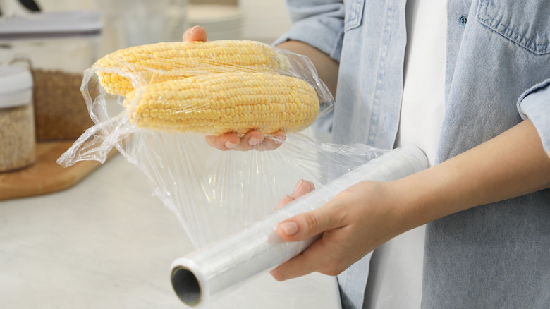 A woman wrapping ears of fresh corn in plastic wrap
