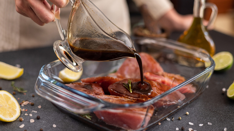 Pouring brown marinade over meat in a glass dish with oil, lemon, and lime pieces on the side.