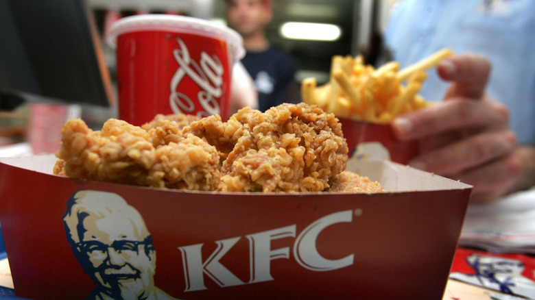 Several pieces of KFC chicken in a cardboard container with a drink and someone grabbing french fries in the background