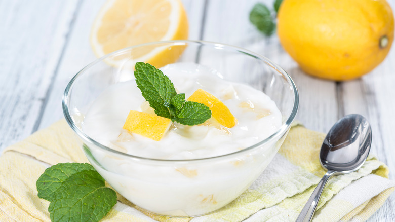 A glass bowl of lemon yogurt on a towel beside a metal spoon with fresh lemons in the background