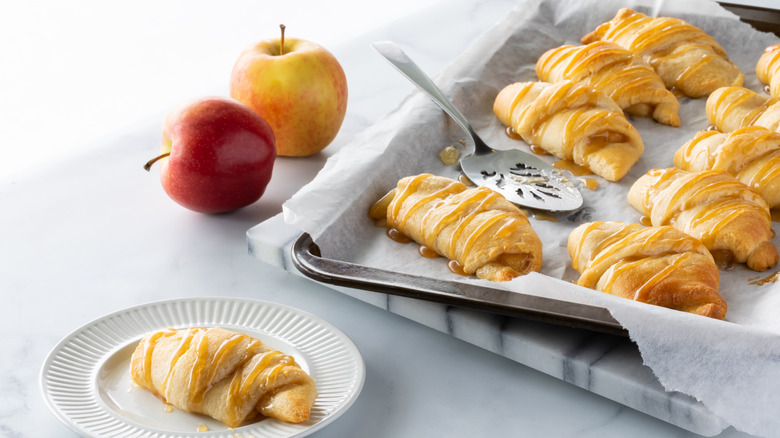 A tray of apple crescent rolls next to one on a plate and two red apples
