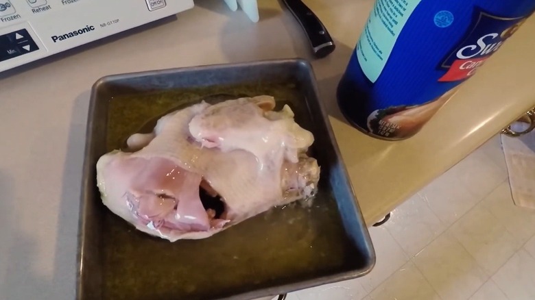 A glistening, pale, whole chicken in a square roasting pan on countertop next to blue can