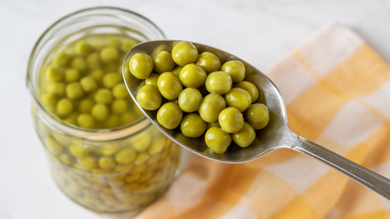 Close up look at a spoon filled with peas being held above a full jar of peas and yellow tea towel.