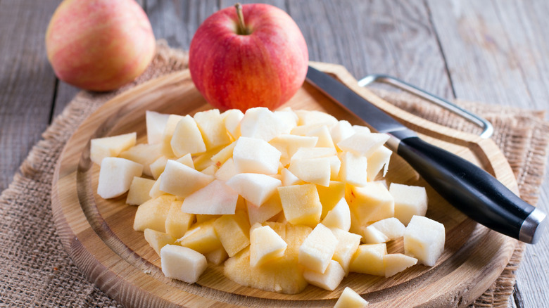 Chopped apples beside a kitchen knife and a whole red apple on a circular wooden cutting board.