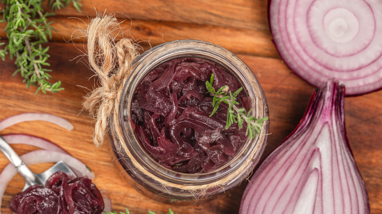 A jar of caramelized red onions sitting on a wooden board with some sliced red onions next to it