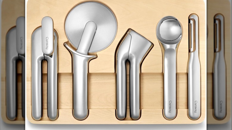 Stainless steel can opener, pizza cutter, garlic press, ice cream scoop, and vegetable peeler in a wooden storage container.