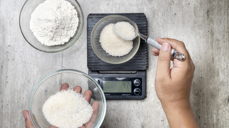 A top view of a person spooning sugar into a bowl resting on a kitchen scale on a rustic grey background