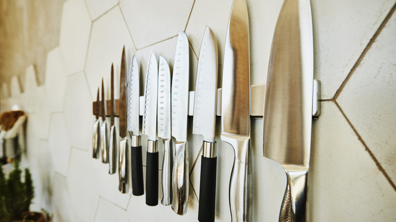 A close-up of assorted knives on a metallic knife strip hanging on a white wall with honeycomb tile