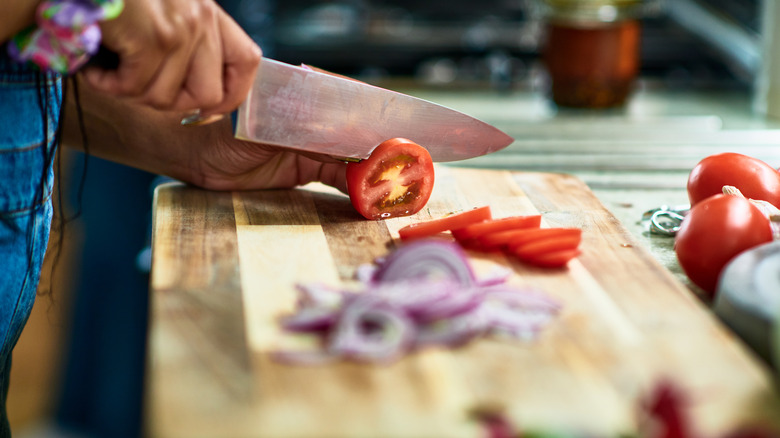 A close-up of a man using a chef's knife to slice tomatos and onions on a wood cutting board