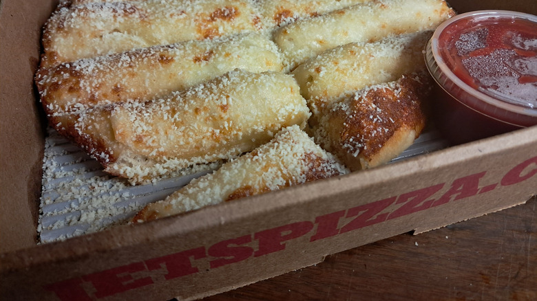Cheesy breadsticks in a cardboard container with a tub of marinara sauce