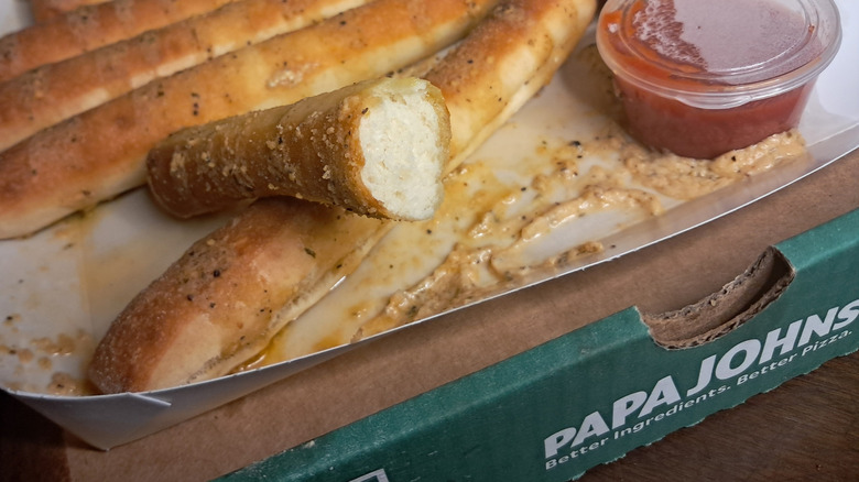 A portion of Papa Johns breadsticks in a cardboard container with a tub of marinara sauce