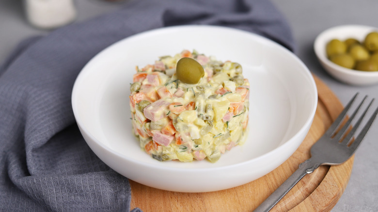 A portion of Ensaladilla Russa garnished with an olive sits in a white bowl on a wooden carving board with a fork and dishcloth next to it, and a bowl of green olives in the background