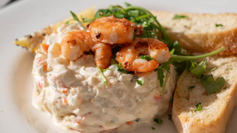 An elaborate portion of Ensaladilla Rusa on a white plate, garnished with shrimp and accompanied by sliced bread