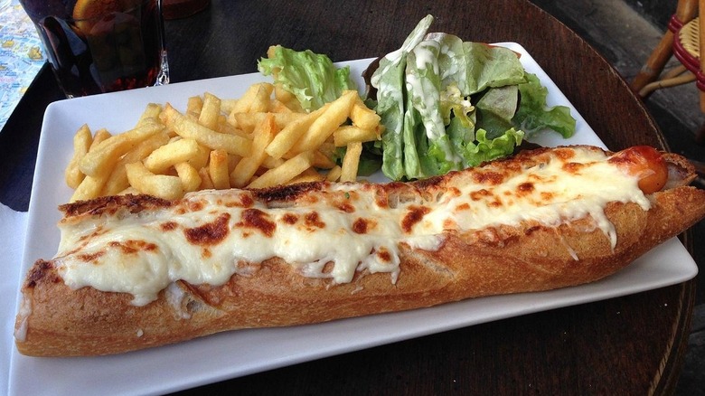 French hot dog in a baguette with melted cheese served on a plate with fries and dressed salad leaves