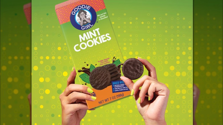 Hand holding a Goodie Girl Mint Cookie up alongside the box on a green and yellow background