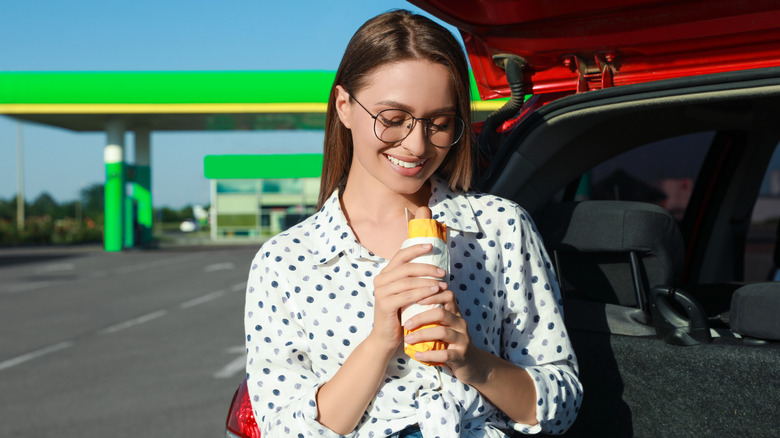 A smiling young woman holds a hot dog while standing by her car in the parking lot of a gas station
