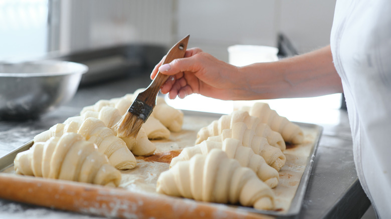 person brushing butter onto croissants on baking sheet
