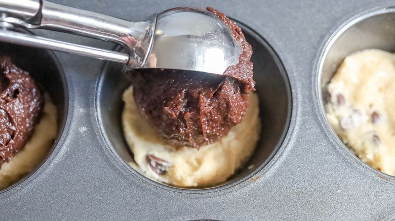 Scoop of brownie batter going into muffin tin well with cookie dough in it