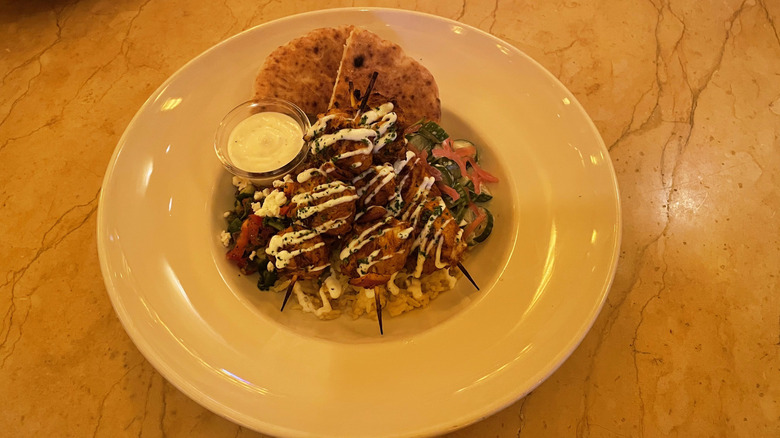 Three chicken skewers sit on top of a bed of rice with tomatoes, cucumbers, and pita bread in a white bowl on a marble table.